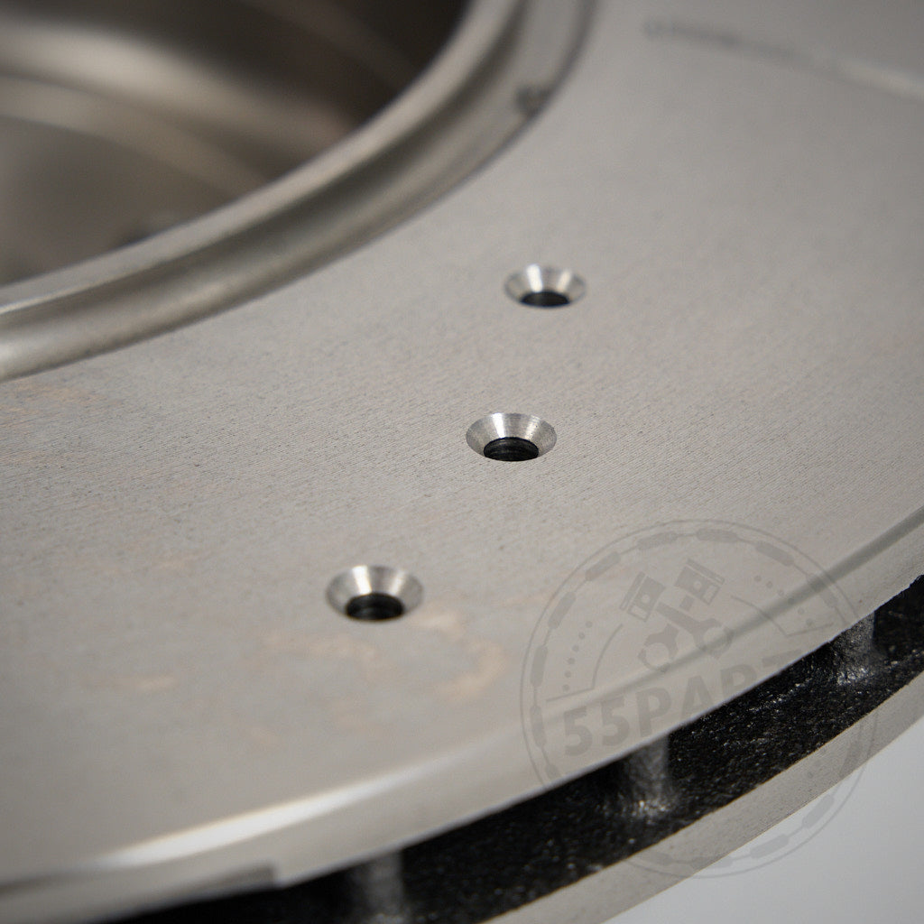 Nahaufnahme eines 55Parts Spezial-Montagesatzes (Bremsscheiben und Buchsen) zur Montage von 2-Kolben-Performance-Bremssätteln an BMW E8x/E9x-Modellen, mit drei Bohrlöchern und einem teilweise sichtbaren Logo im Hintergrund.