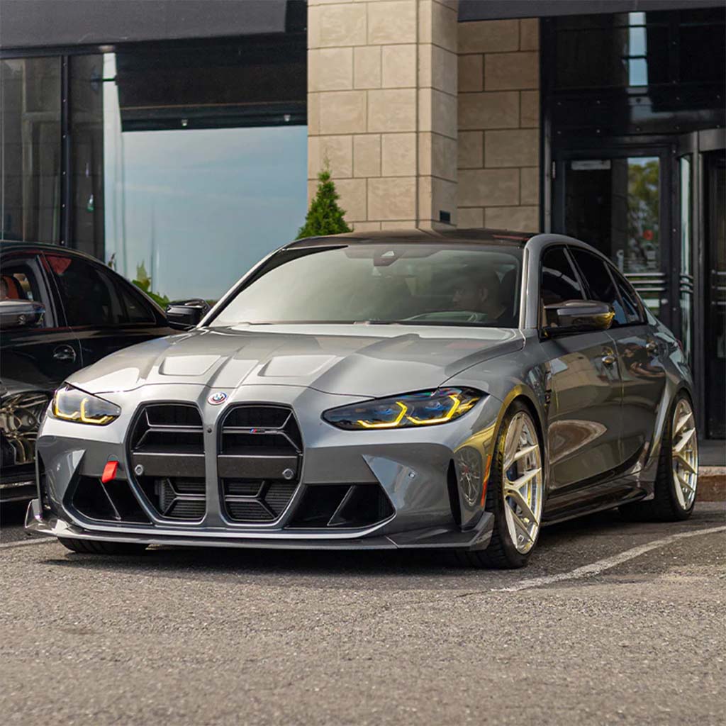 Ein grauer BMW mit gelben Scheinwerfern und einem Twistnfix TNF+ CSL Style Carbon Grill für BMW M3/M4 (G80/G81/G82/G83) incl. Teilegutachten ist vor einem modernen Gebäudeeingang geparkt.