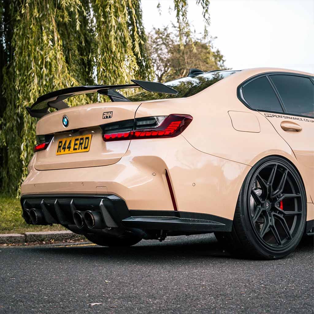 Ein beiger BMW M3 mit schwarzen Felgen und einem Twistnfix TNF+ Performance Heckflügel Carbon parkte am Straßenrand unter einem belaubten Baum.