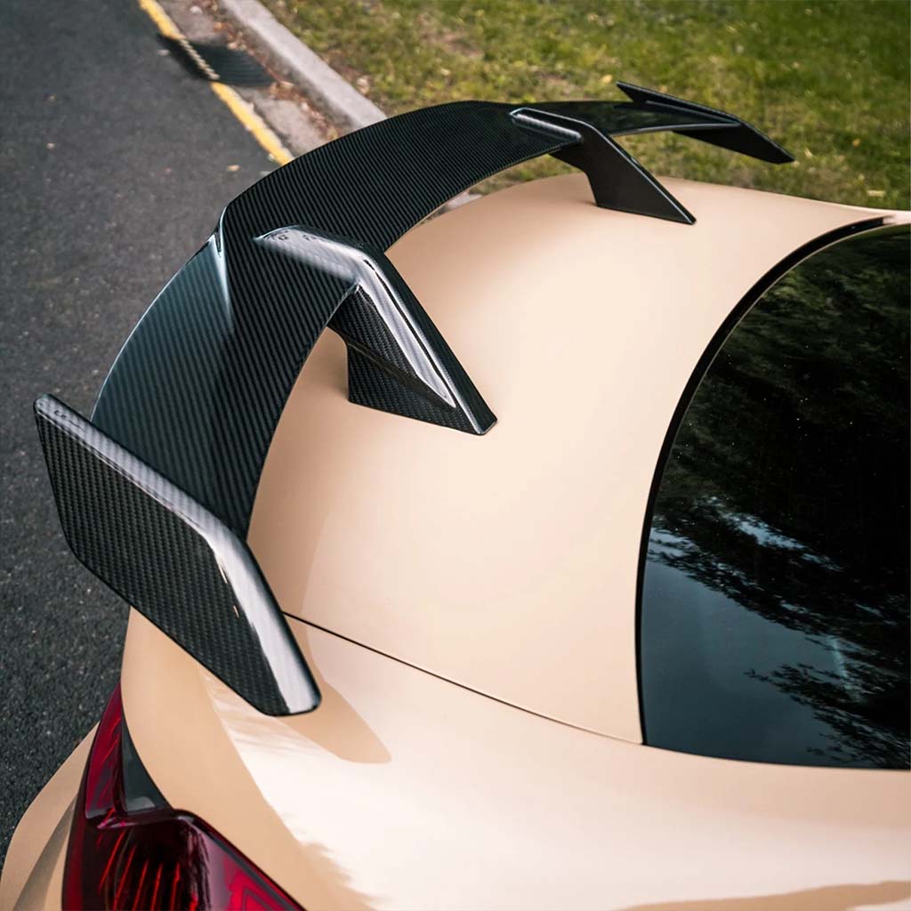 Nahaufnahme eines beigen Autos mit einem Twistnfix TNF+ Performance Heckflügel Carbon für BMW M3 (G80), M4 (G82), oder M2 (G87), inklusive Teilegutachten, geparkt neben einem Bordstein und einer Grasfläche.