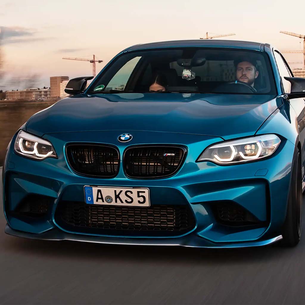 Ein blauer BMW M2 mit einer ZAERO Design EVO-S Frontlippe für F87 N55 fährt auf einer Straße, zwei Personen darin, Stadtgebäude im Hintergrund.