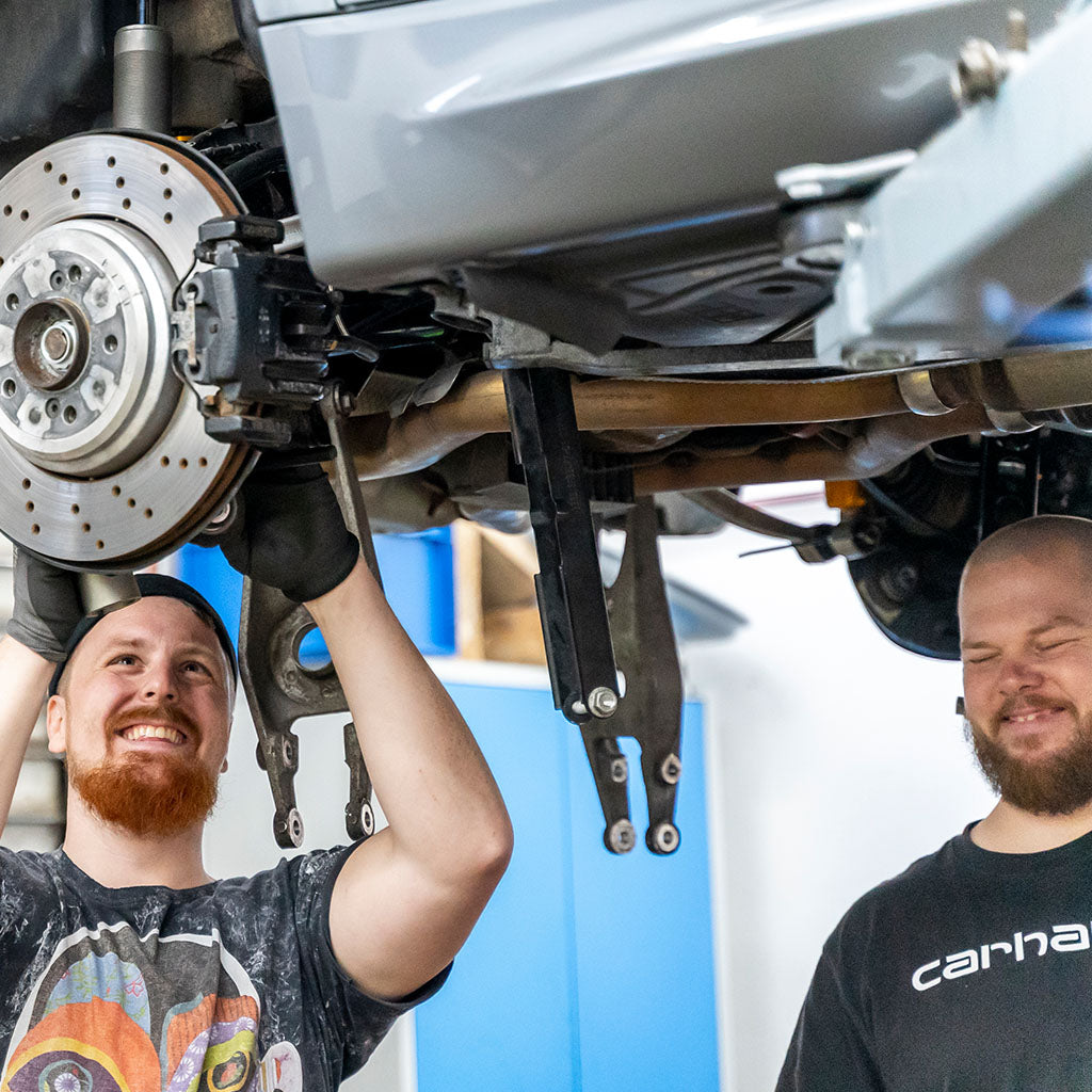 Zwei lächelnde Mechaniker arbeiten unter einem angehobenen Auto und überprüfen das Bremssystem in einer hellen Autowerkstatt mit Qualitätsteilen.