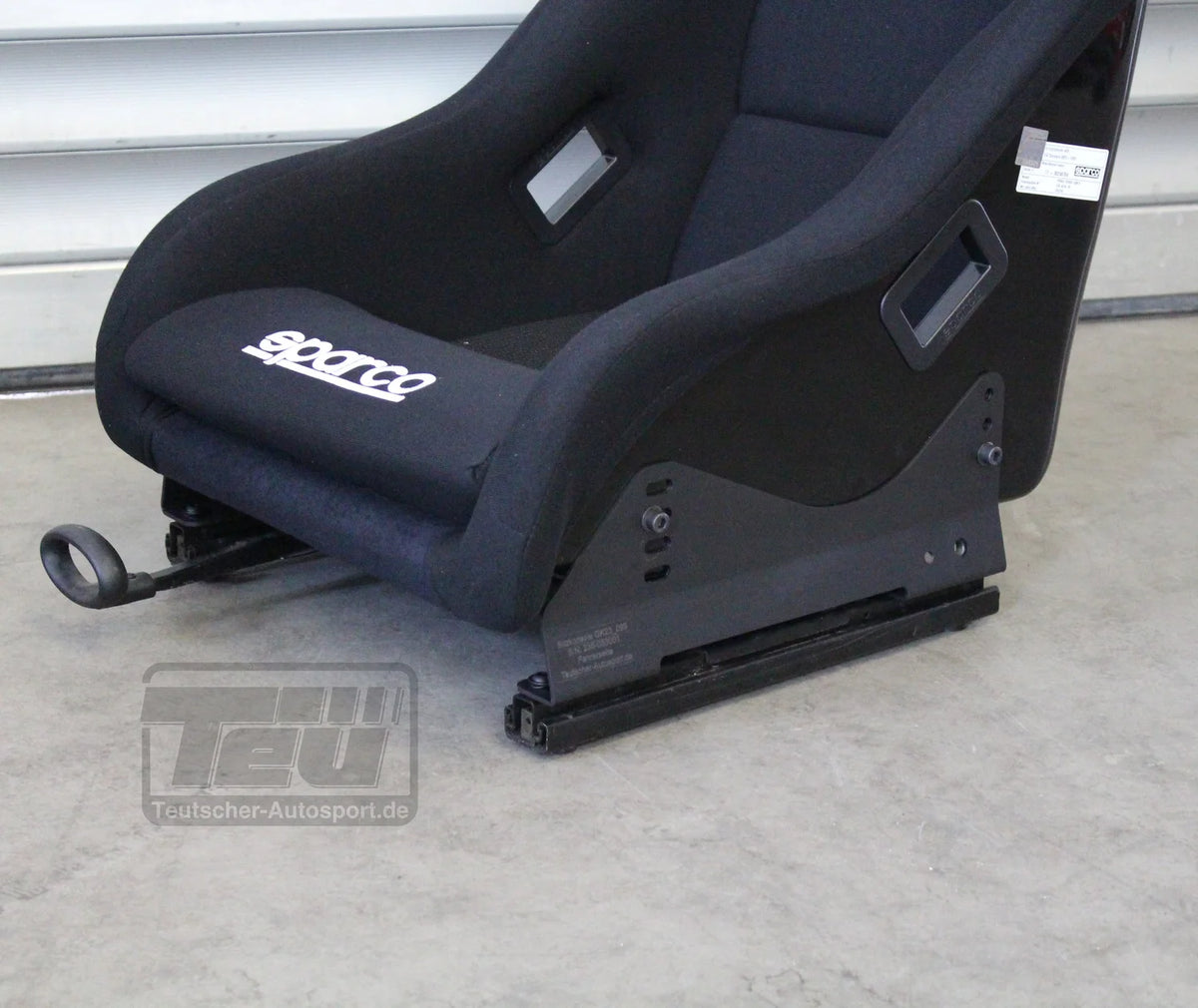 Schwarzer Sparco Rennsitz mit Teutscher Recaro, Sparco, OMP Sitzkonsolen für Mini R50/R53 inkl. Materialnachweis, steht auf einem Betonboden vor einer Metallwand.