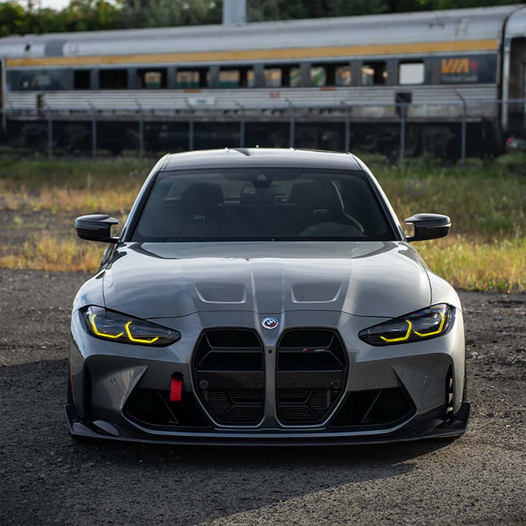 Ein grauer BMW Sportwagen mit gelben Scheinwerfern und einem Twistnfix TNF+ CSL Style Carbon Grill für M3 (G80/G81) oder M4 (G82/G83) ist vor einem stehenden Zug geparkt.