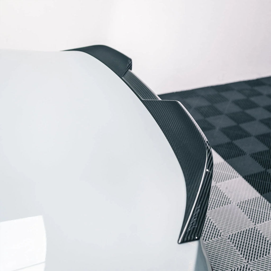 Nahaufnahme des Twistnfix TNF+ Performance Carbon Ducktail Spoilers für BMW M2 (G87)/2er (G42) mit Teilegutachten, montiert an einem weißen Auto; schwarz-grau karierter Boden im Hintergrund sichtbar.