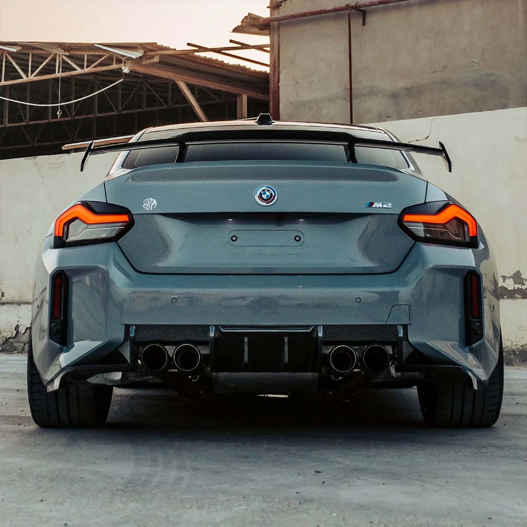 Heckansicht eines grauen BMW M2 (G87) mit Twistnfix MHC TNF+ Edition 1 Heckdiffusor Carbon, inklusive Teilegutachten, vor einem verwitterten Gebäude geparkt und mit vier Auspuffrohren versehen.