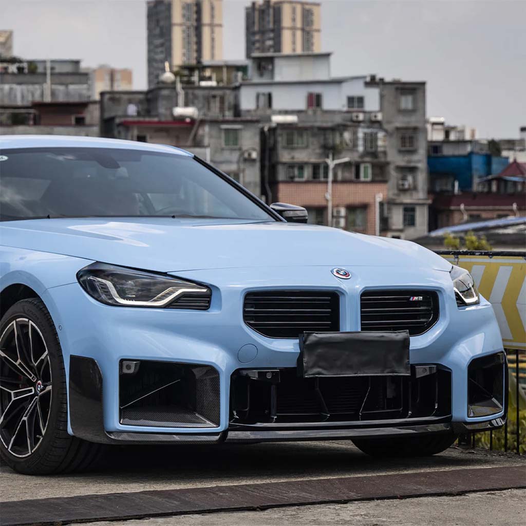 Ein hellblauer BMW M2 (G87) mit einem Twistnfix TNF+ Performance Frontspoiler Carbon inkl. Teilgutachten. Teilegutachten wird von städtischen Gebäuden geparkt, teilweise sichtbar von der vorderen linken Seite.