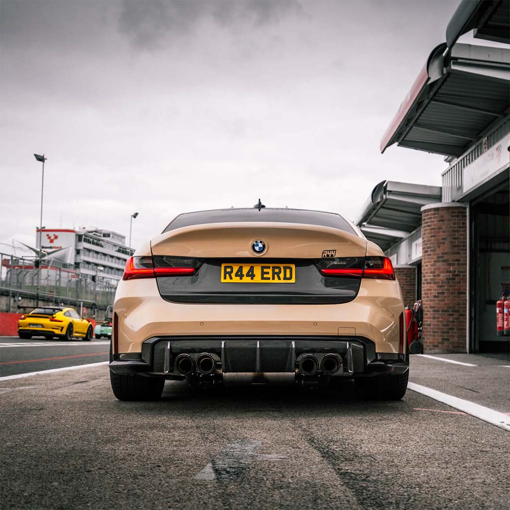 Ein bronzener BMW mit dem Kennzeichen R44 ERD steht auf einer Rennstrecke, ausgestattet mit dem Twistnfix TNF+ Performance Heckdiffusor Carbon für M3 (G80/G81) und M4 (G82/G83); im Hintergrund ist ein gelbes Auto zu sehen.