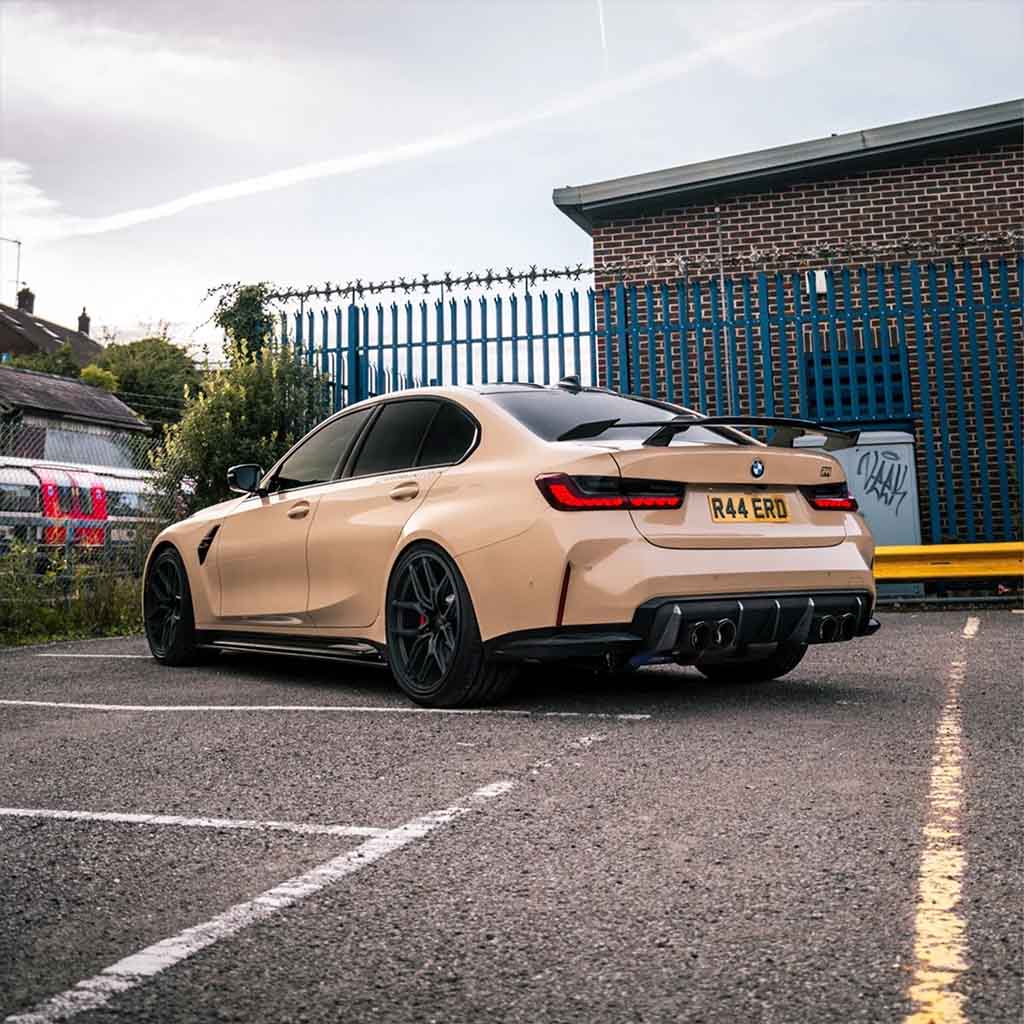Ein beiger BMW M3 (G80) mit schwarzen Akzenten und einem Twistnfix TNF+ Performance Heckflügel Carbon ist auf einem städtischen Parkplatz geparkt, von hinten gesehen, und unterstreicht sein G8X-Plattformdesign.