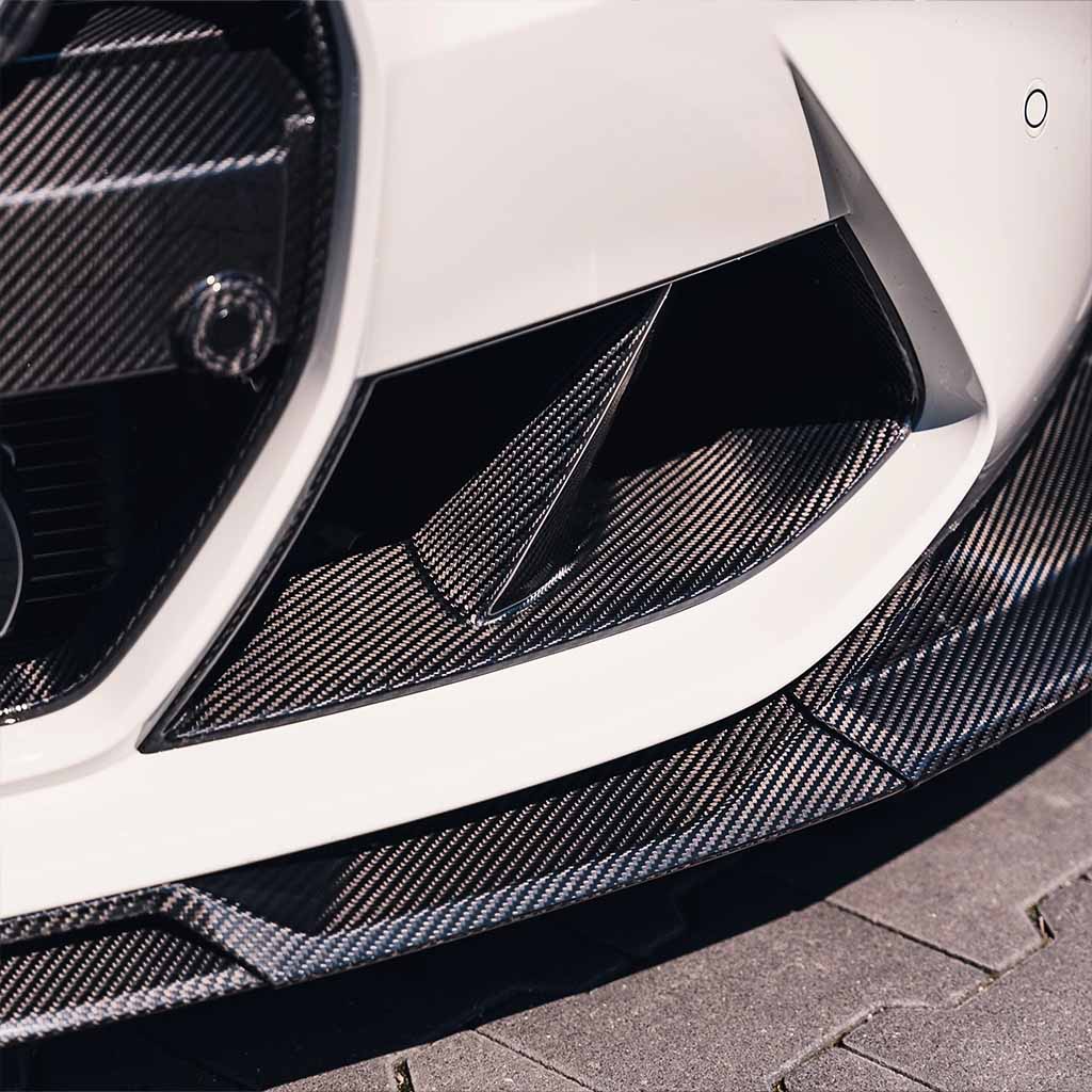Nahaufnahme der Frontstoßstange eines weißen Fahrzeugs mit Twistnfix TNF+ Performance Lufteinlässe Carbon für BMW M3 (G80/G81) & M4 (G82/G83), mit Prepreg-Carbon-Details und sportlichem Design, inkl. Teilegutachten.