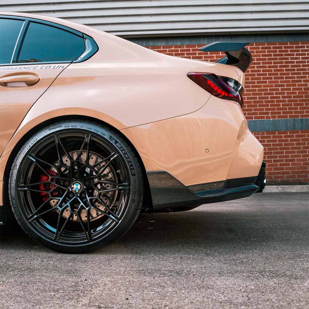 Nahaufnahme eines beigen Sportwagens mit Twistnfix TNF+ Performance Seitendiffusoren Carbon für BMW M3 (G80/G81) mit Teilegutachten an Hinterrad, Spoiler und Rückleuchten vor einer Backsteinwand.