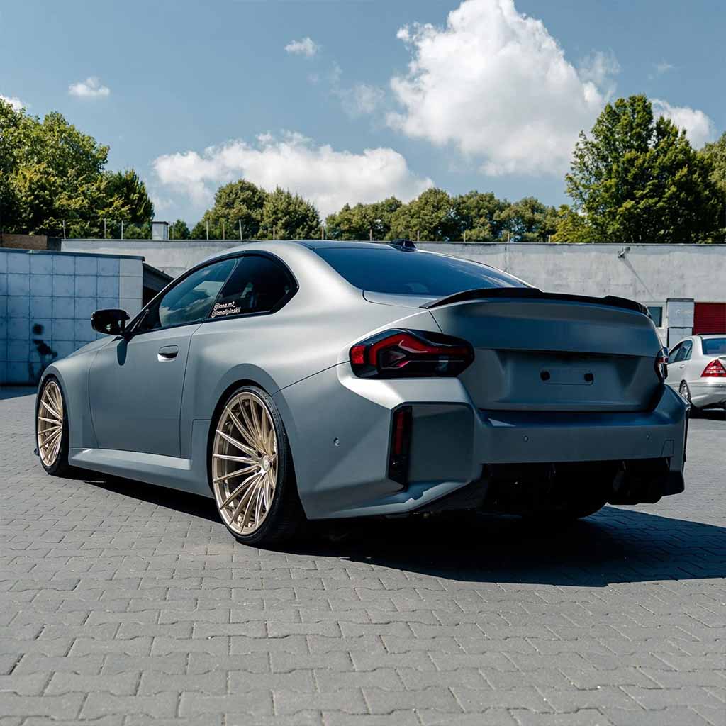 Mattgrauer Sportwagen mit goldenen Felgen und einem Twistnfix TNF+ V-Style Ducktail für BMW M2 (G87)/2er (G42) mit Teilgutachten im OEM-Plus-Look, geparkt auf einem gepflasterten Parkplatz bei leicht bewölktem Himmel.