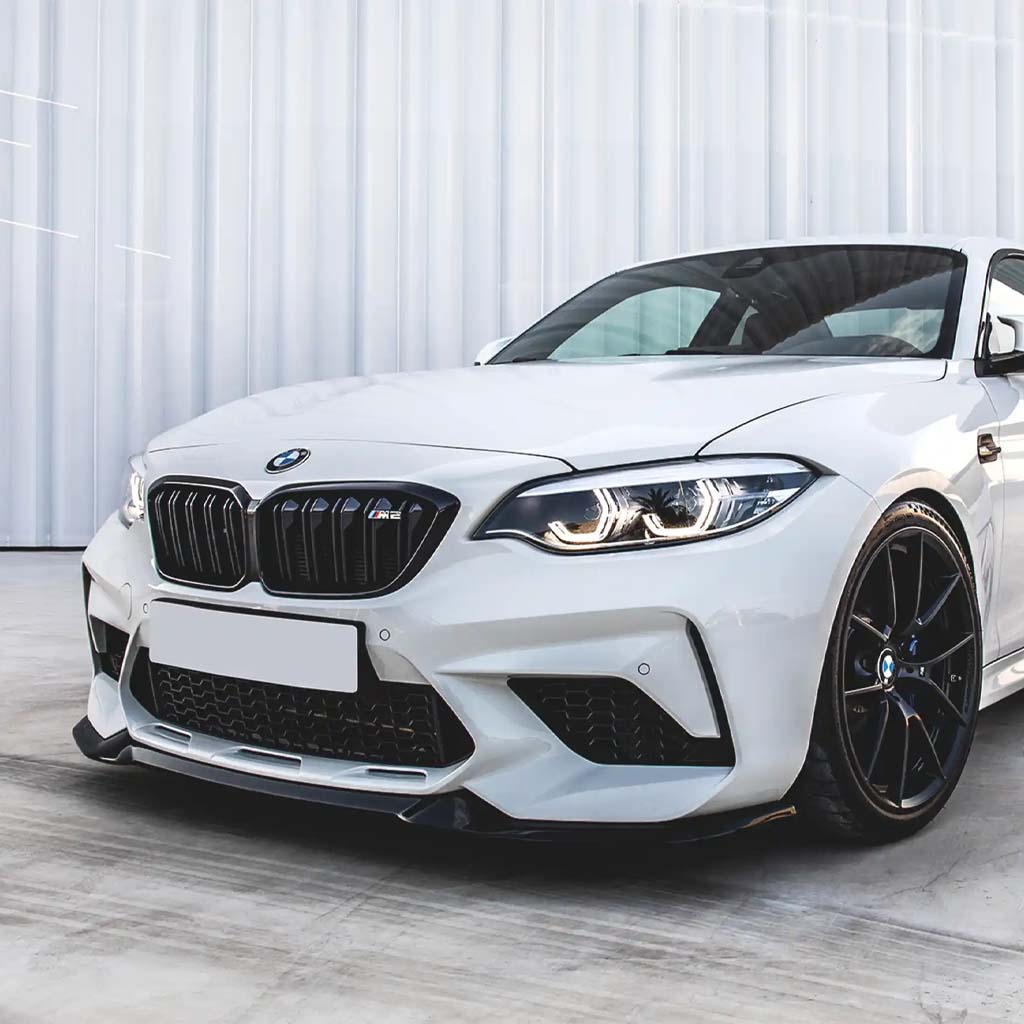 Ein weißer BMW F87 Competition mit einer ZAERO Design EVO-S Frontlippe ist auf einer Betonfläche vor einem modernen weißen Gebäude geparkt.