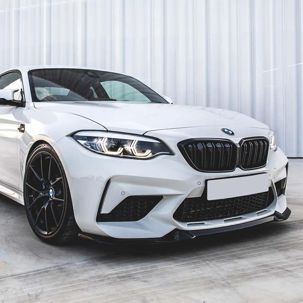 Ein weißer BMW F87 Competition mit einer ZAERO Design EVO-S Frontlippe ist auf einer Betonfläche vor einem modernen weißen Gebäude geparkt.