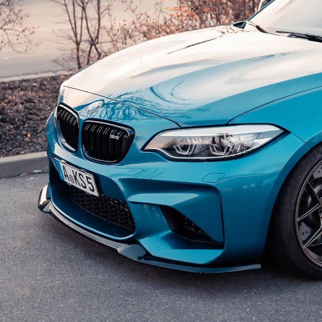 Ein blauer BMW M2 mit einer ZAERO Design EVO-S Frontlippe für F87 N55 fährt auf einer Straße, zwei Personen darin, Stadtgebäude im Hintergrund.