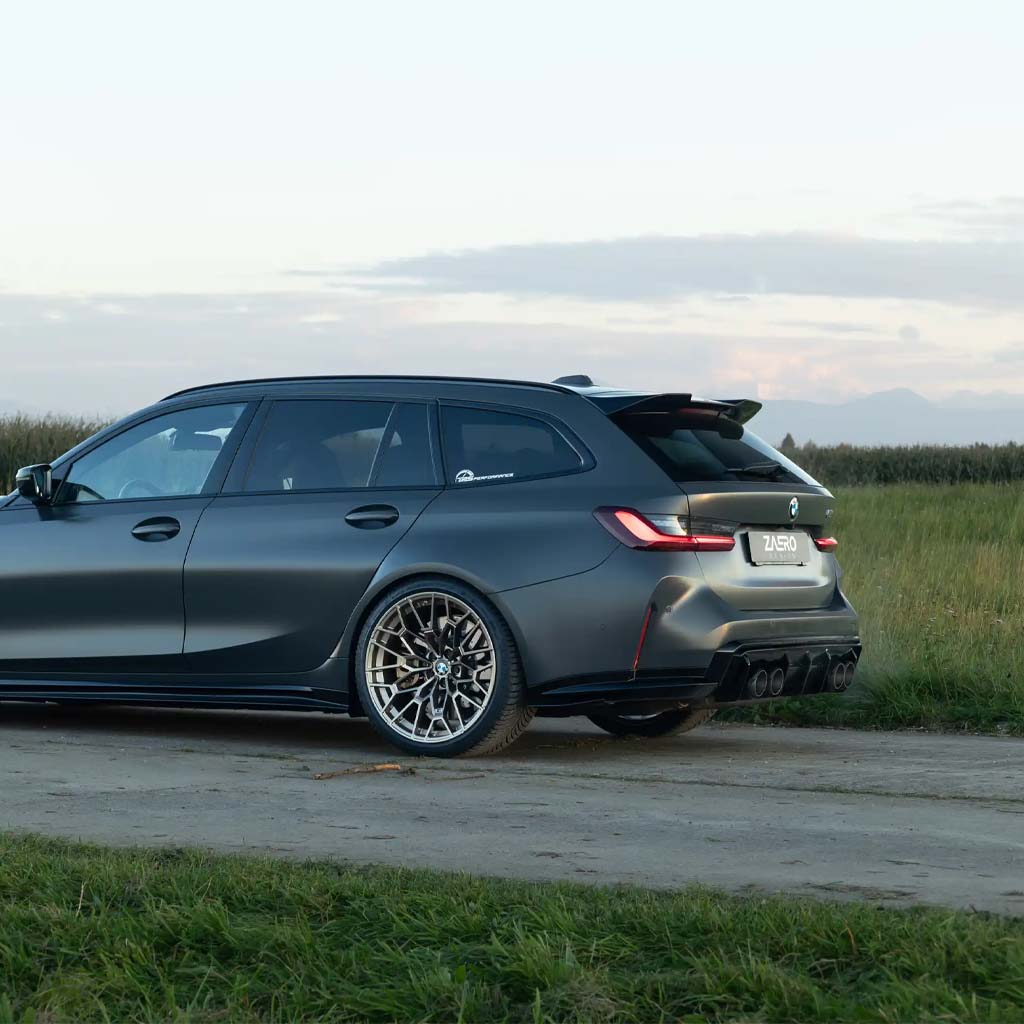 Ein mattschwarzer BMW Kombi mit einem ZAERO Design EVO-S Heckspoiler, der für BMW M3 G81 und G21 entwickelt wurde, ist auf einem Feldweg neben einer Wiese unter einem bewölkten Himmel geparkt.