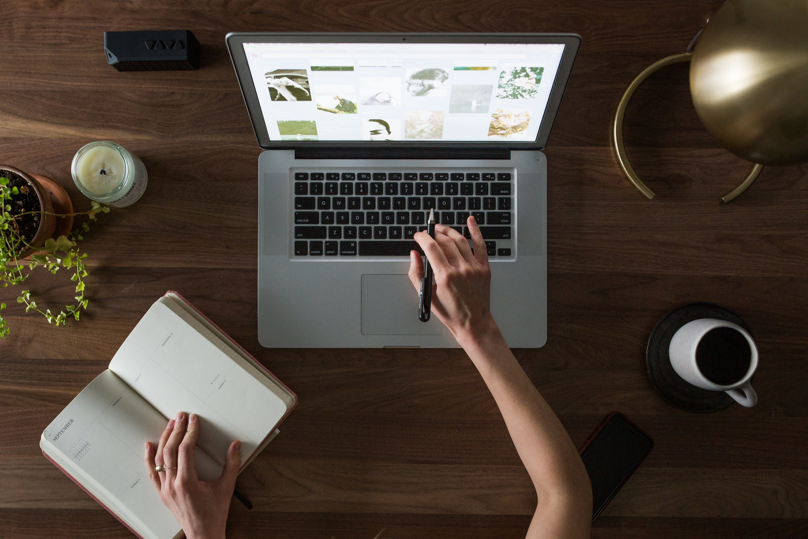 Person, die einen Laptop verwendet und in ein Notizbuch an einem Holzschreibtisch mit Kaffee, Pflanzen, Kerze und natürlichem Licht schreibt.