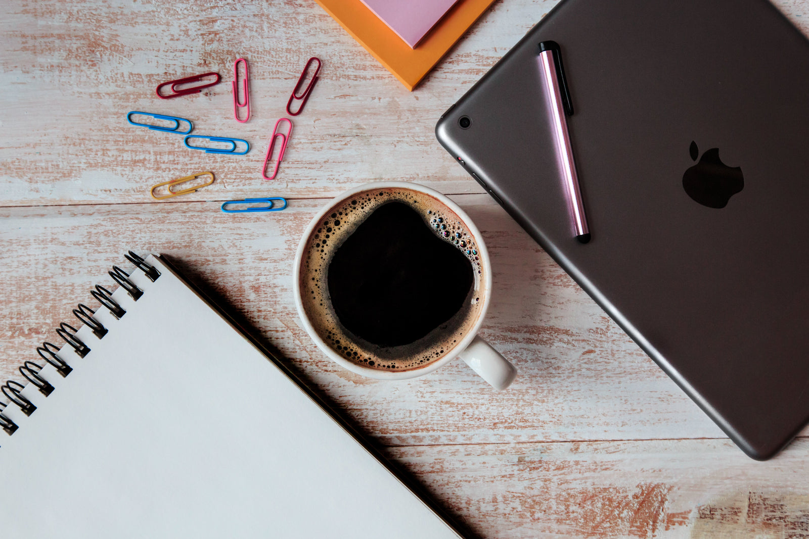 Tasse Kaffee, leerer Notizblock, bunte Büroklammern, iPad mit Stift und Haftnotizen auf einem Holzschreibtisch – perfekt, um Ihren Arbeitsplatz zu organisieren.