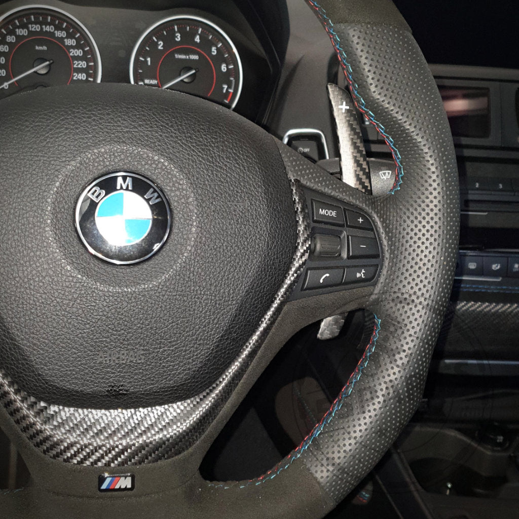 Nahaufnahme der 55Parts Aluminium/Carbon Schaltwippen Paddles, passend für BMW 1/235 und 40i, 3/435 und 40i, M2, M3 und M4 F-Serie, abgebildet mit Mode-Tasten und bunten Nähten am Lenkrad.