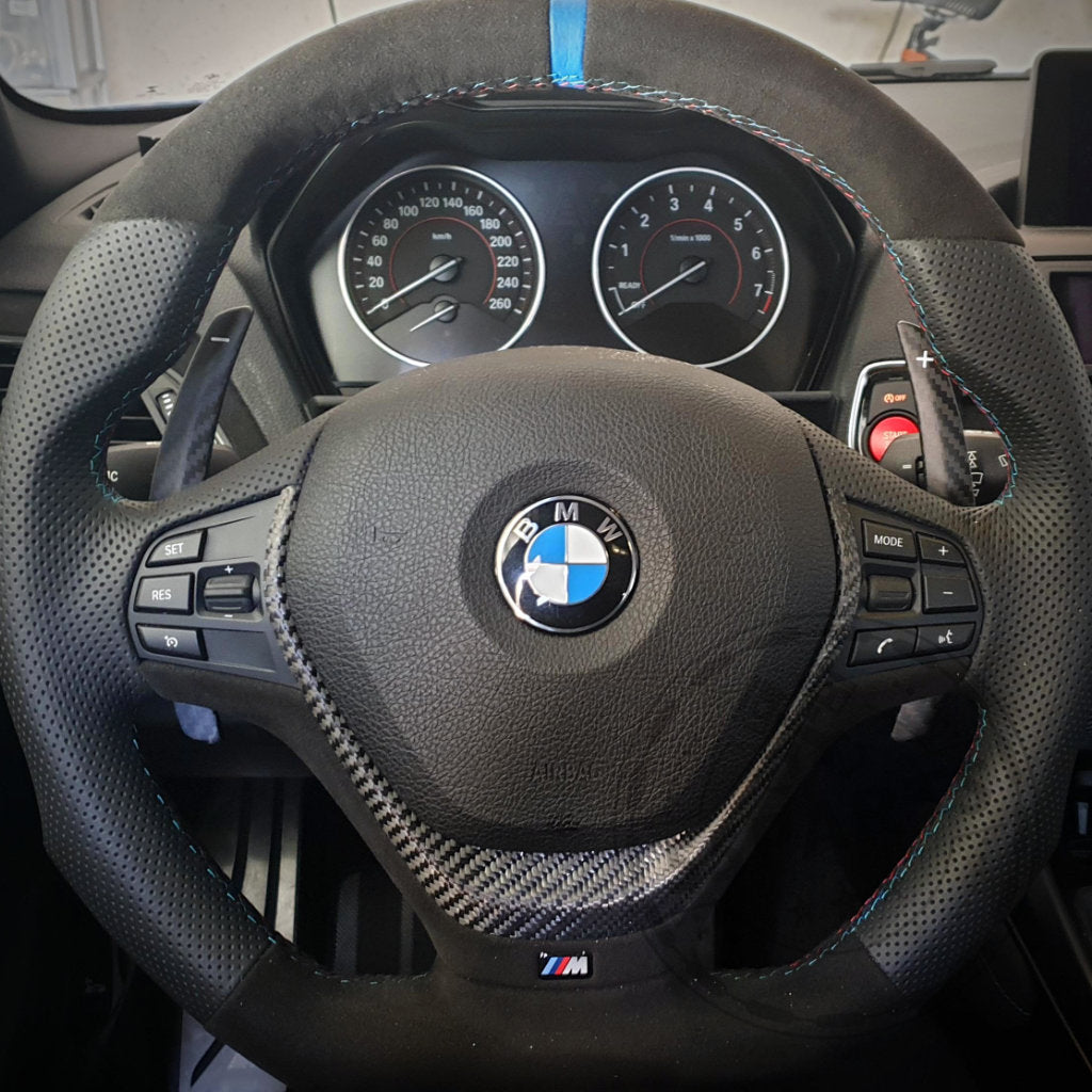Nahaufnahme eines BMW F-Serie Lenkrads mit 55Parts Aluminium/Carbon Schaltwippen für 1/235, 3/435, 40i, M2, M3 &amp; M4 und sichtbaren Armaturenbrettanzeigen und Bedienknöpfen.