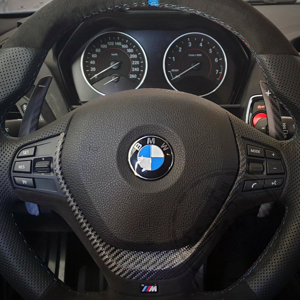 Nahaufnahme eines BMW F-Serie Lenkrads mit 55Parts Aluminium/Carbon Schaltwippen Paddles passend für BMW 1/235 und 40i, 3/435 und 40i, M2, M3 und M4 F-Serie, sowie Armaturenbrettanzeigen und Bedientasten.